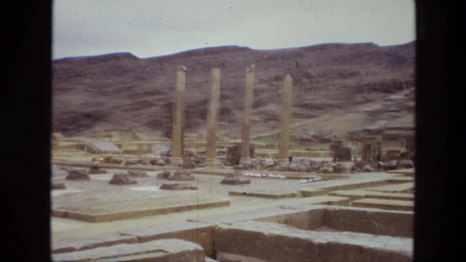 1972 Iran 8mm frame showing a wider view of columns, stepped paving, foundations, and a hillside beyond the ruins.