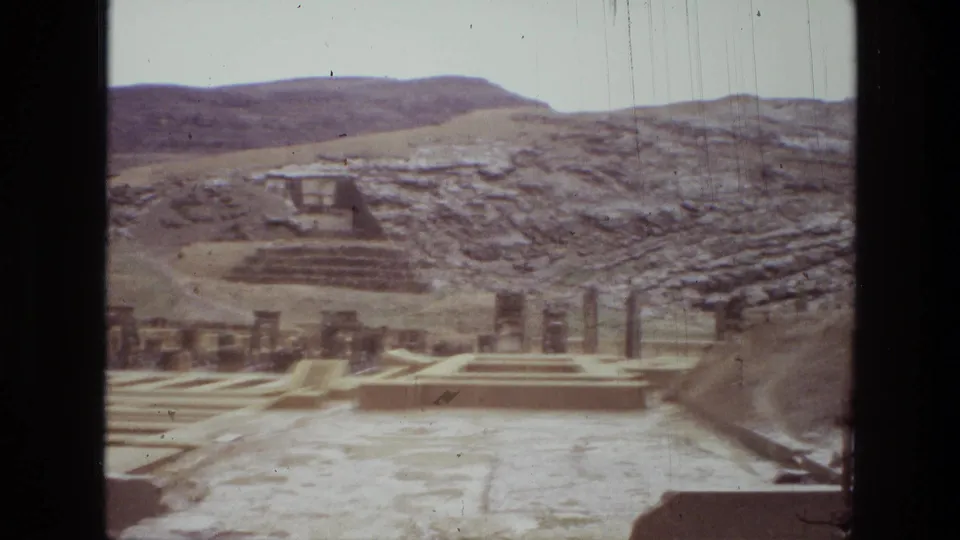 1972 8mm frame of Persepolis ruins in Iran, showing columns, terraced stone areas, and arid mountains behind the complex.