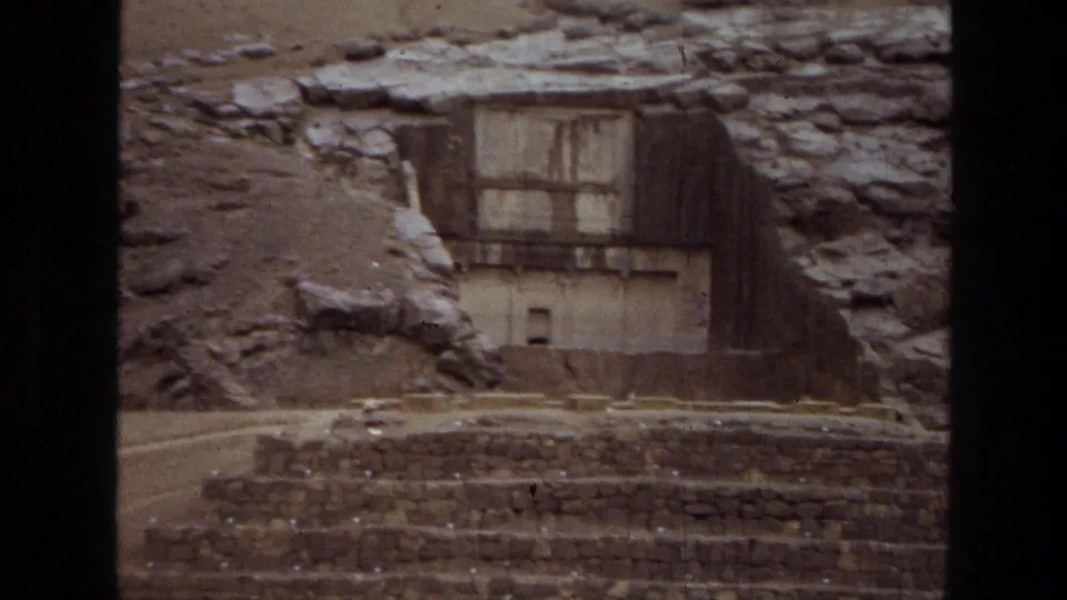 1972 8mm frame from Iran showing a rock-cut stone structure set into a hillside above weathered stepped platforms.