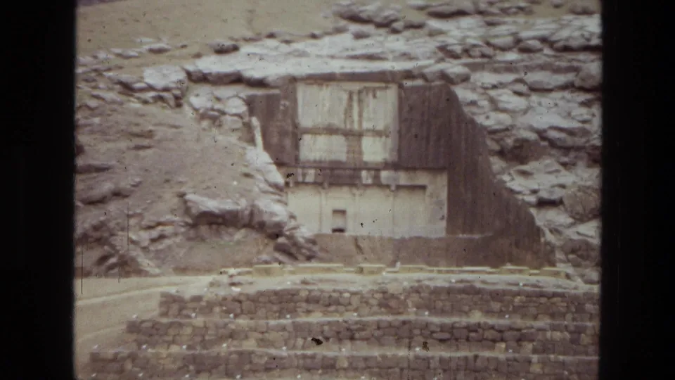 1972 Iran travel footage frame showing a weathered stone facade, hillside rock, and stepped masonry in the foreground.