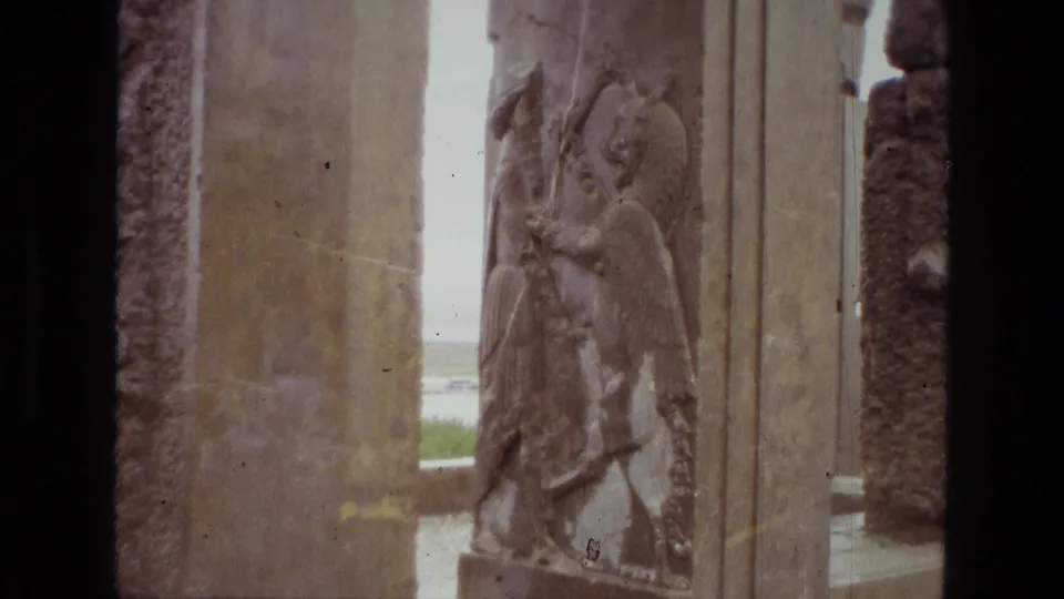 1972 Iran 8mm frame showing weathered stone pillars and a bas-relief winged figure at an archaeological site.