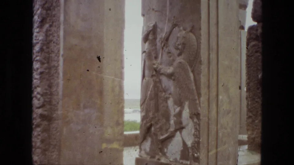 1972 Iran home-movie view of a carved stone relief beside massive ancient pillars, with grass and sky in the background.