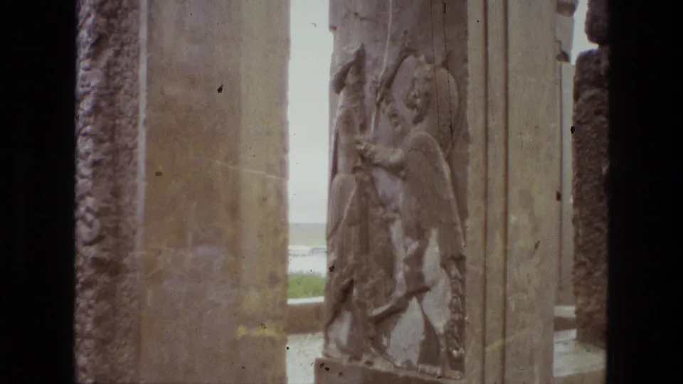 1972 Iran 8mm close framing of a winged bas-relief figure on weathered stone, filmed from a visitor's path through ruins.