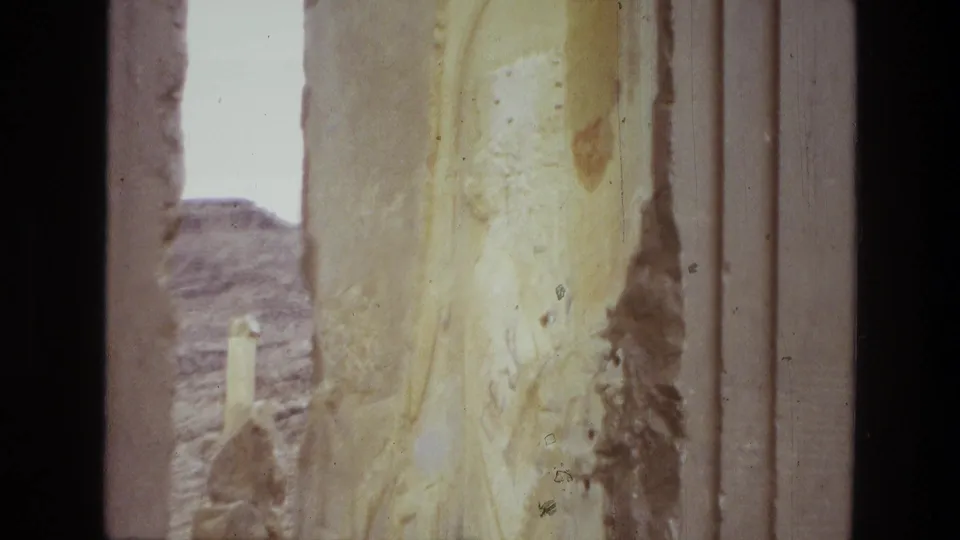 A faded 8mm frame from Iran shows close stone carving, pillar edges, and a distant rocky backdrop through the ruins.