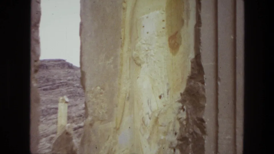 1972 Iran 8mm frame showing a close weathered stone column at a ruin site with pale sky and rocky desert hills behind it.