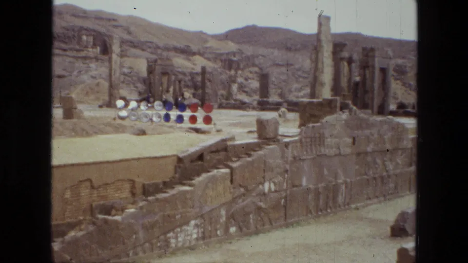1972 Iran 8mm view of a ruin site with standing columns, low stone walls, sandy ground, hills, and red, blue, and white circular objects.