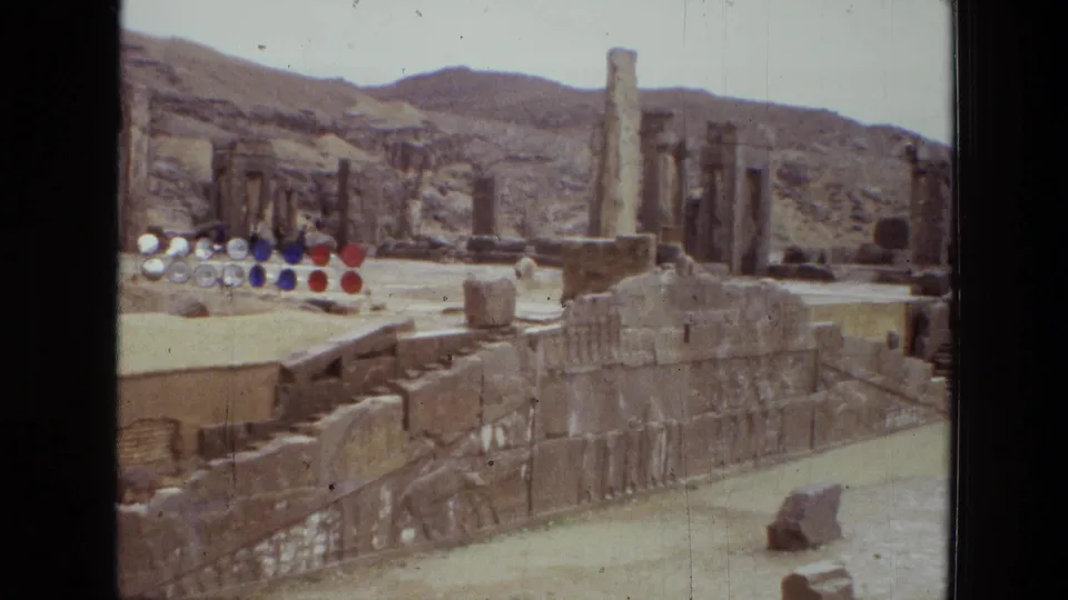 1972 Iran 8mm frame showing a wide visitor view of ancient stone ruins with columns, stepped platforms, sandy ground, and hills beyond.