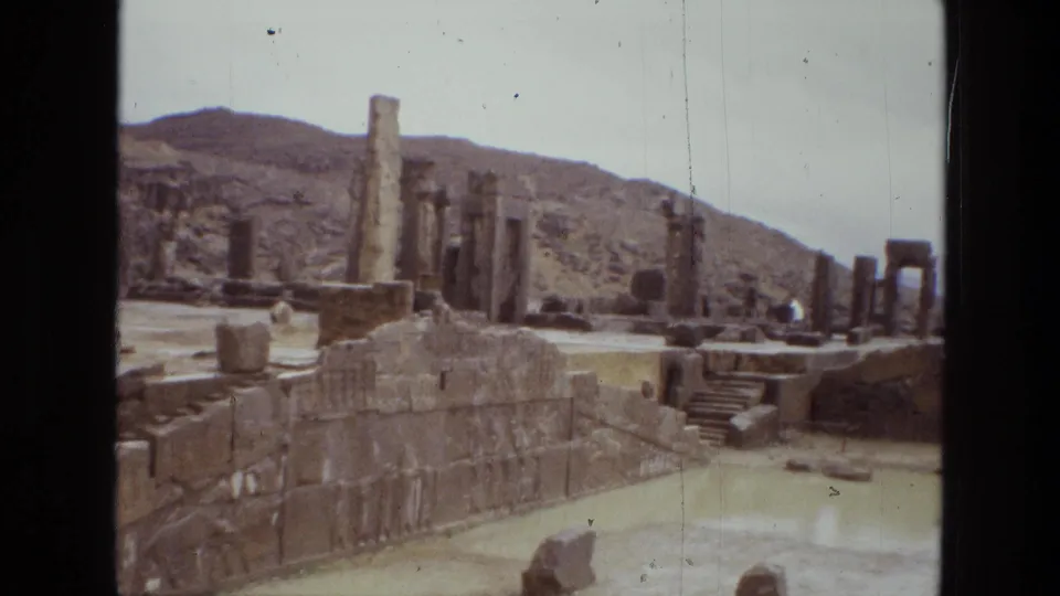 1972 Iran 8mm frame showing a stone platform, upright columns, broken walls, steps, sandy foreground, and distant desert hills.