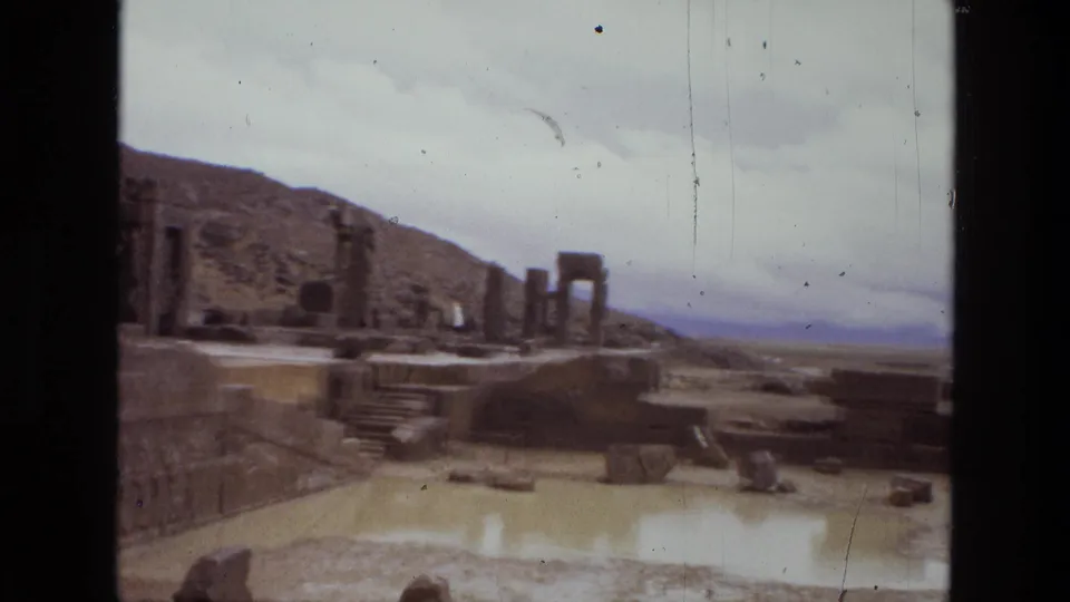 Handheld 1972 Iran travel footage frames ruined stone platforms, columns, steps, and broken blocks against a barren hillside.