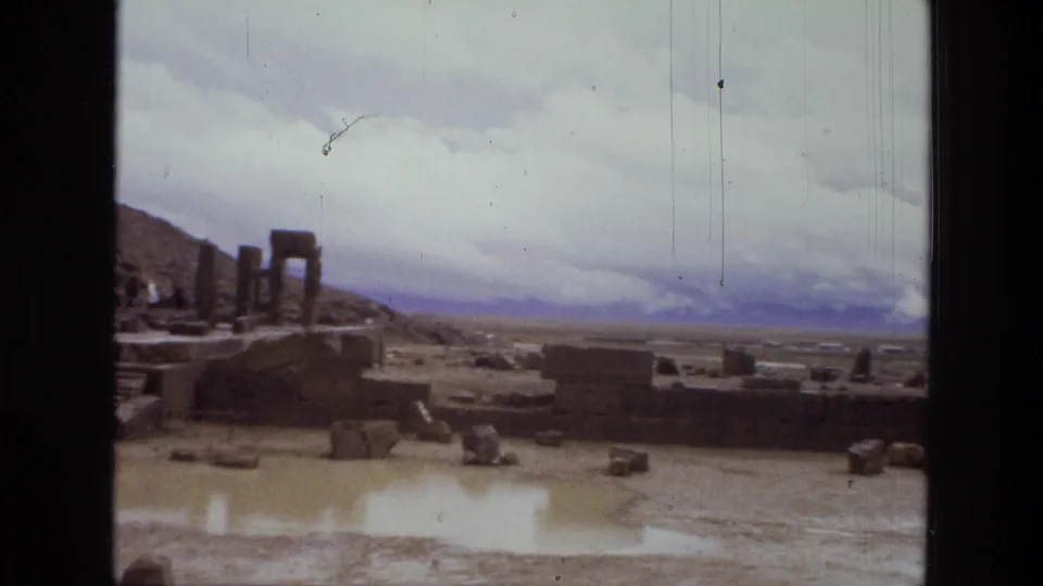 A 1972 8mm frame from Iran shows the ruins receding toward a broad valley, with columns at left and a muddy pool below.
