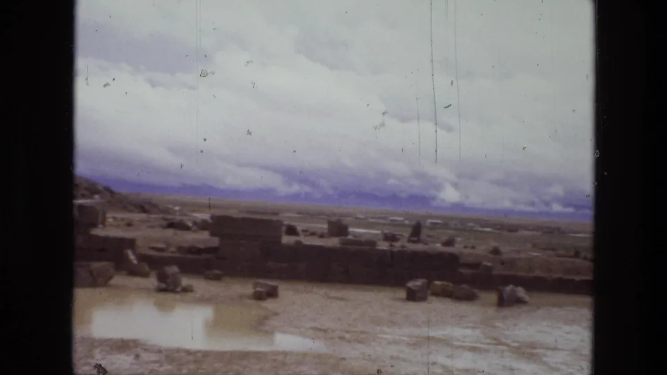 Final 1972 Iran frame shows lower stone remains, scattered blocks, wet ground, and distant mountains beneath a cloudy sky.