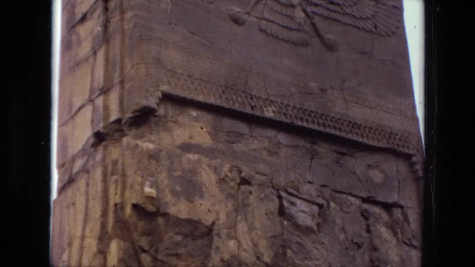 1972 8mm frame from Iran showing a tall weathered stone surface with a winged emblem and eroded bas-relief details.