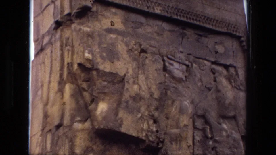 1972 Iran home-movie frame showing eroded stone relief work, missing sections, and a partial winged motif on the carved surface.