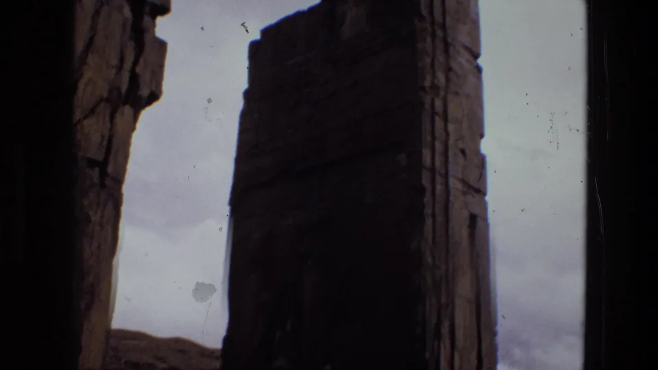 1972 Iran 8mm frame with a tall, thick stone column or slab rising against a hazy sky, with rough ruin surfaces nearby.