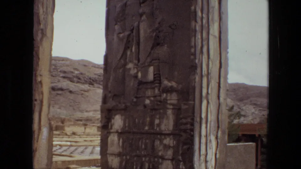 1972 Iran 8mm frame showing a carved upright stone column near an open ruin area, with low hills and a small structure in the distance.