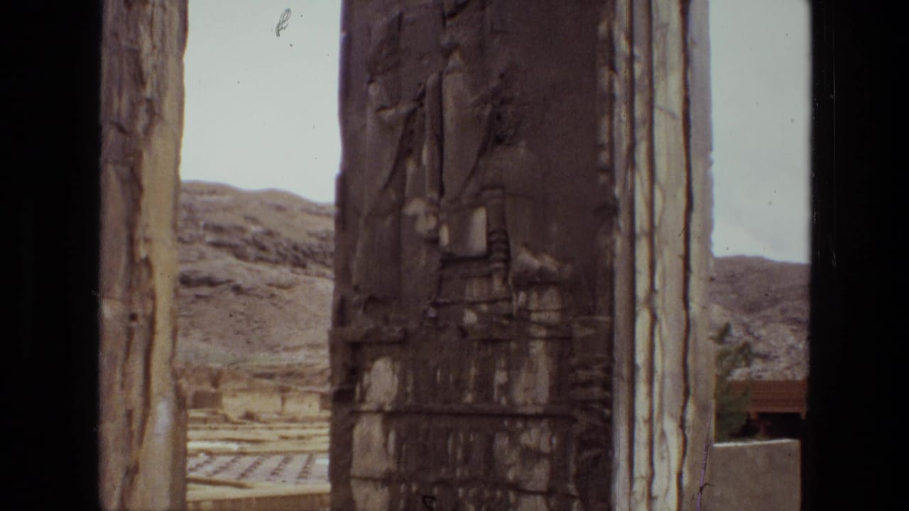 1972 Iran home-movie frame of an ancient carved stone pillar, stone uprights, pale sky, and dry hills beyond the archaeological site.
