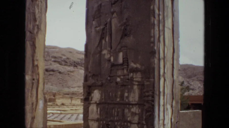 1972 Iran home-movie frame of an ancient carved stone pillar, stone uprights, pale sky, and dry hills beyond the archaeological site.