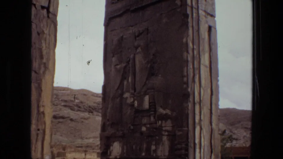 1972 Iran 8mm still with a darker close view of carved stone relief, a vertical opening, pale sky, and rocky hills in the background.