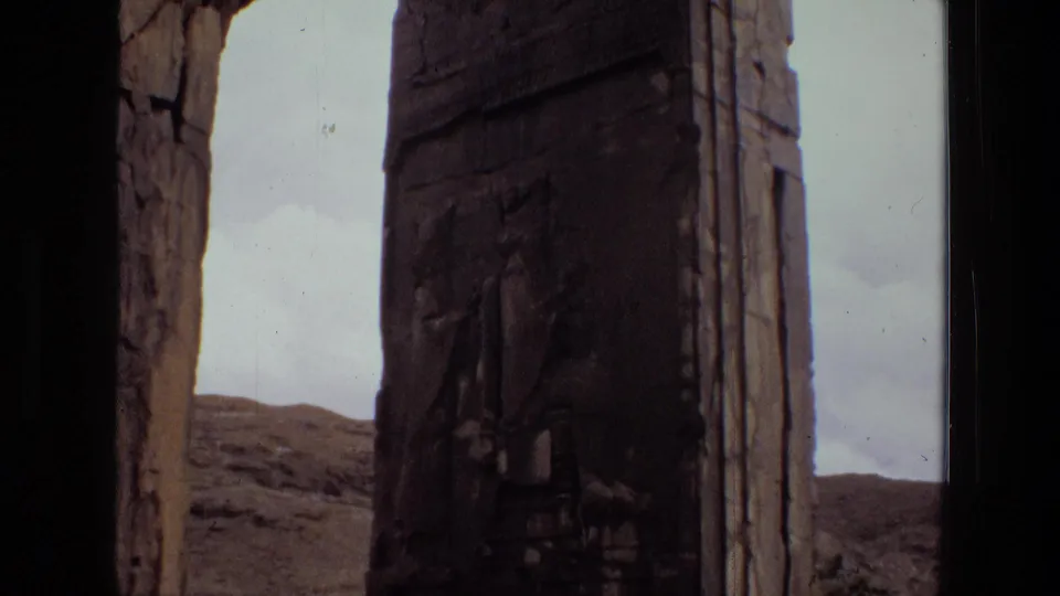 1972 Iran archival frame showing a tall weathered carved pillar in close view, with stonework edges, pale sky, and arid hills beyond.