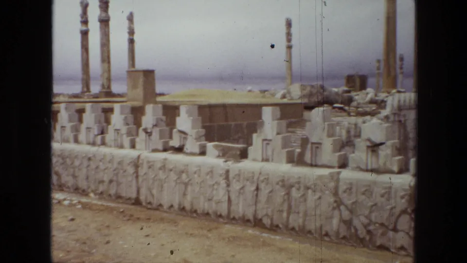 1972 8mm view across an Iranian ruin site with bas-relief figures along a wall, upright columns, and loose architectural stones.