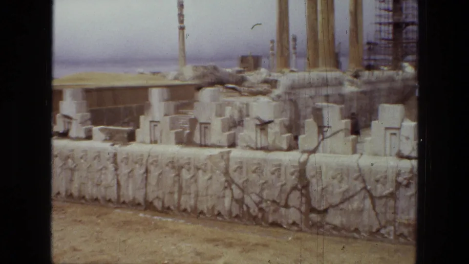 1972 8mm frame from Iran showing carved reliefs, standing columns, stacked stones, and scaffolding within an ancient ruin complex.