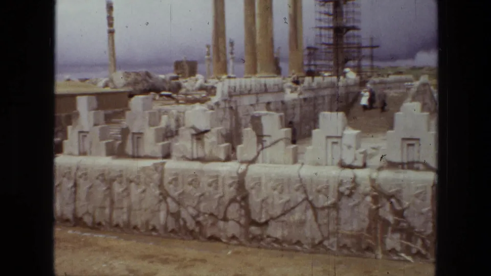 1972 8mm view of Persepolis in Iran, showing carved stone reliefs, block ruins, tall columns, and scaffolding in the background.