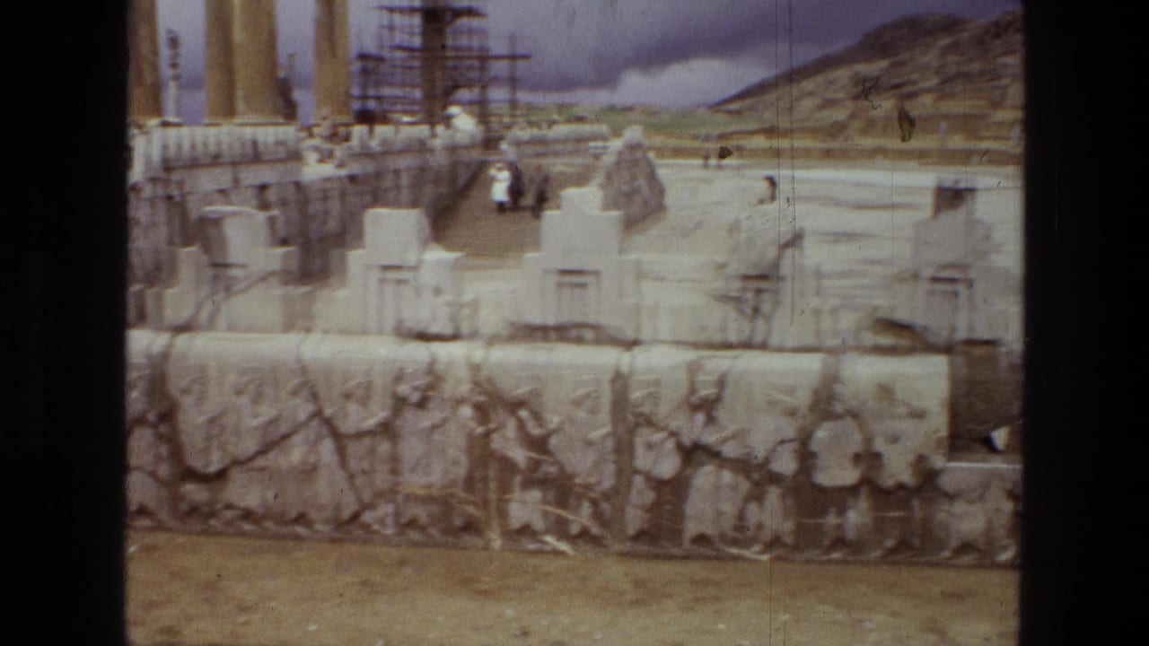 1972 8mm frame of Persepolis ruins in Iran, with carved reliefs, reconstructed stone forms, columns, scaffolding, and small distant visitors.