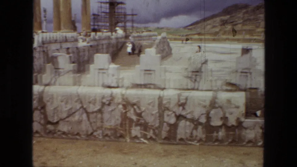 1972 8mm frame of Persepolis ruins in Iran, with carved reliefs, reconstructed stone forms, columns, scaffolding, and small distant visitors.