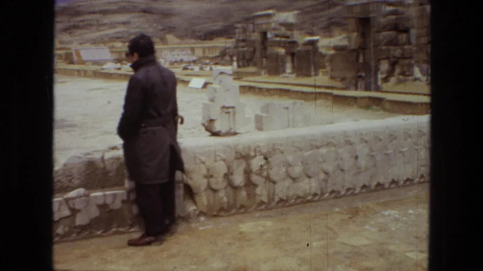 Visitor-shot 1972 8mm view at Persepolis in Iran, with a man in a dark coat beside carved stone reliefs and column ruins.