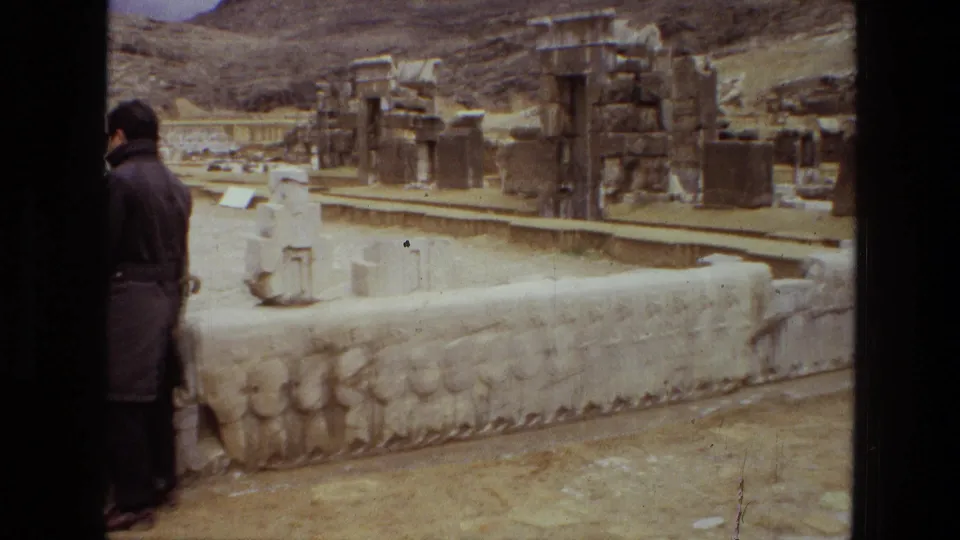 1972 8mm frame from Persepolis, Iran, showing a man beside a carved relief wall with broken columns, ruins, and hillside terrain beyond.