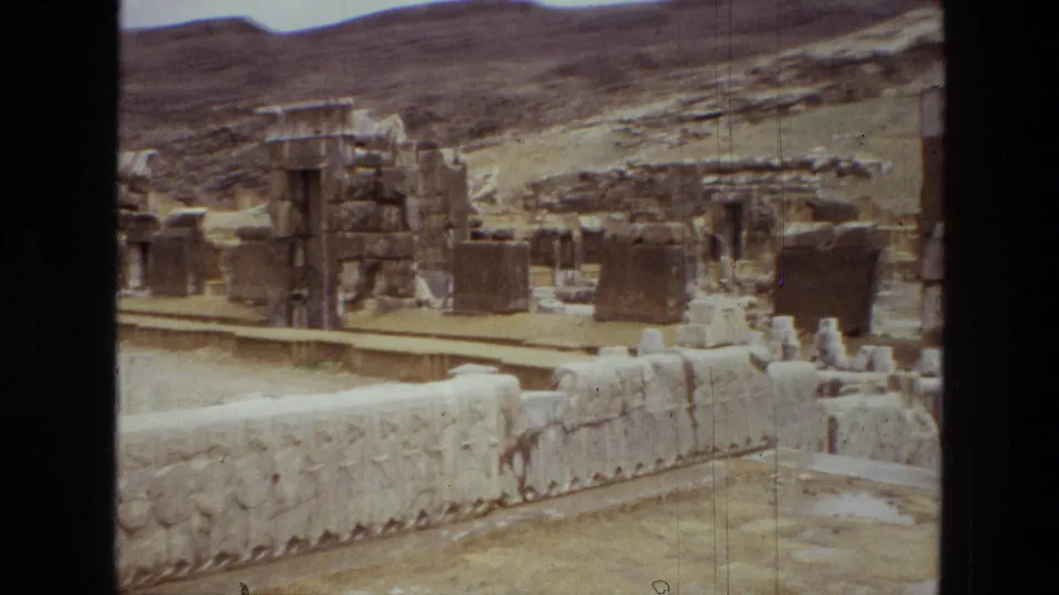 1972 Iran 8mm frame with weathered stone structures, a relief-lined wall, and open hillside beyond.