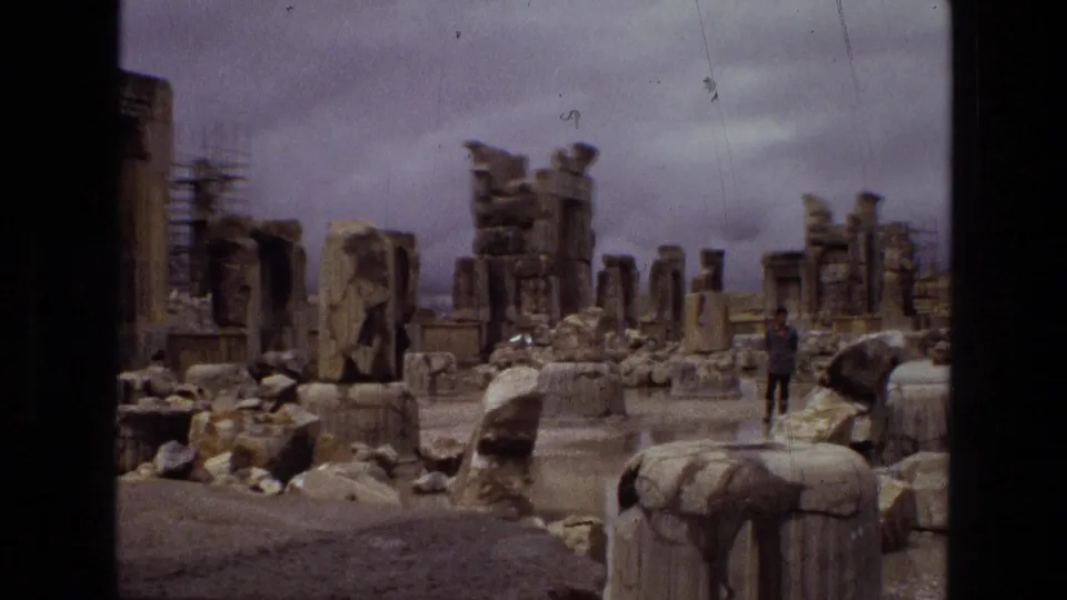 1972 Iran frame showing a wide visitor-shot view of stone ruins, broken columns, scattered rubble, and a distant person under a cloudy sky.