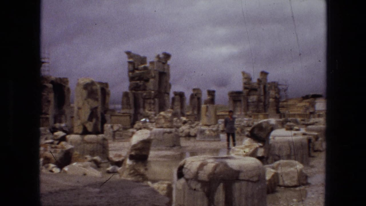 1972 Iran 8mm frame showing ancient ruins, collapsed columns, scattered stone blocks, wet pavement, and a distant tourist under gray clouds.