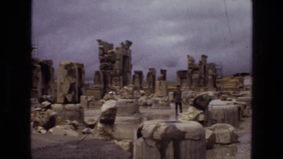 1972 Iran 8mm frame showing ancient ruins, collapsed columns, scattered stone blocks, wet pavement, and a distant tourist under gray clouds.
