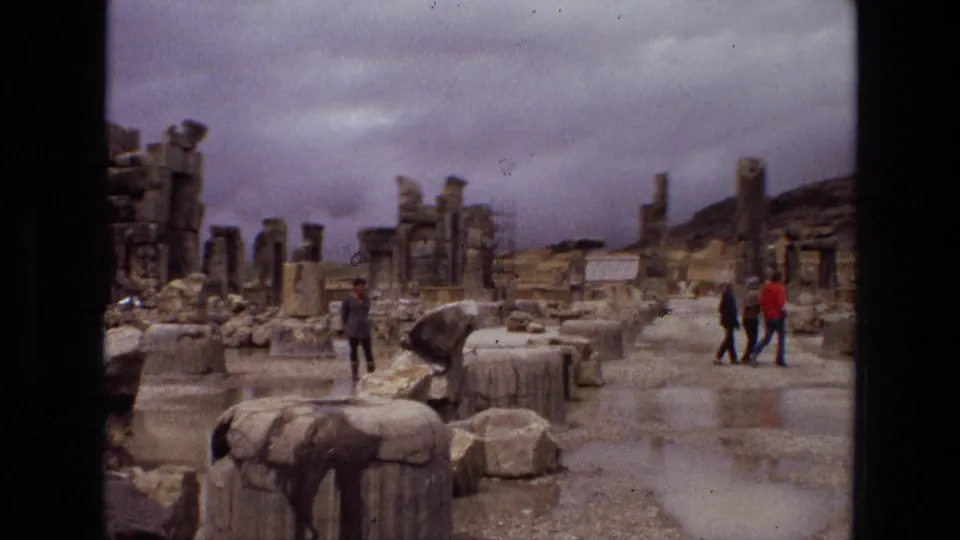 1972 Iran travel footage frame with tourists walking among ancient stone ruins, broken columns, rubble, and reflective rainwater.