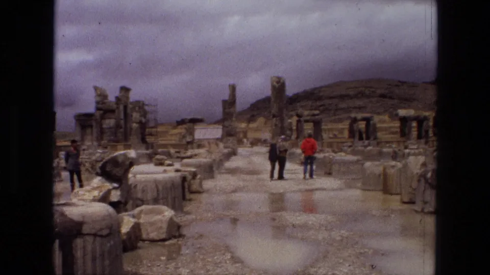 1972 Iran 8mm frame showing tourists crossing a rain-slick archaeological site with broken pillars, rubble, and hillside ruins beyond.