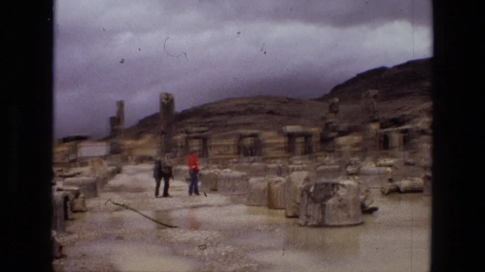 1972 Iran home-movie frame with two visitors near scattered ancient stonework, rain-darkened ground, gray sky, and a low hillside behind the ruins.