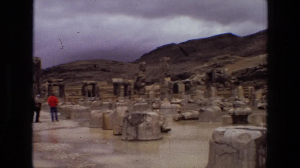 1972 8mm footage from Iran showing distant visitors among scattered stone ruins, columns, and architectural fragments under an overcast sky.