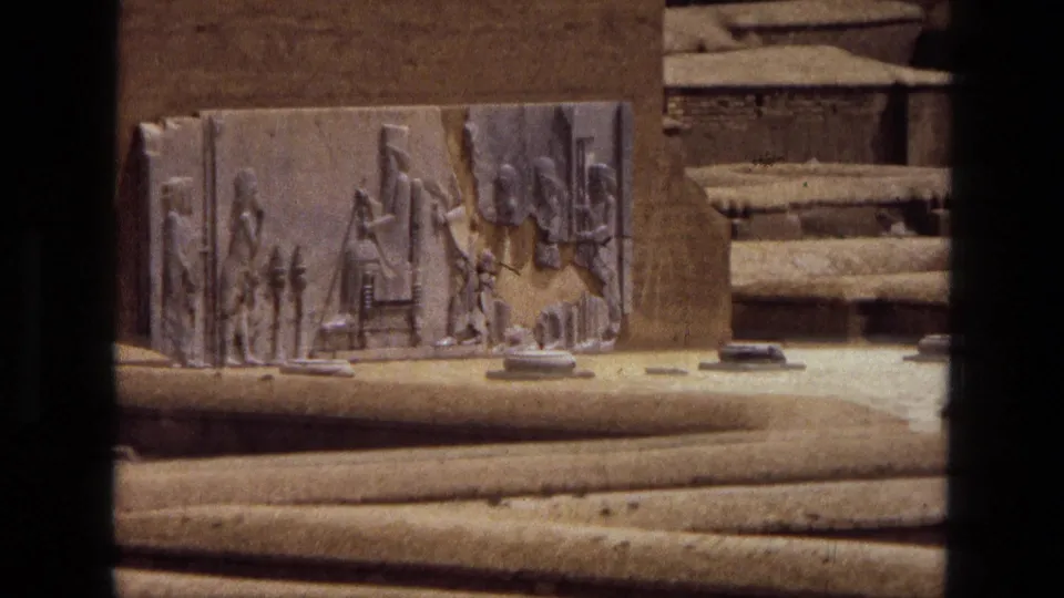 1972 Iran 8mm frame showing a carved relief slab on a sandy exterior, partly masked by dark film or foreground borders.