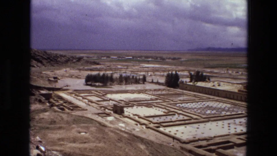 1972 Iran 8mm frame showing a broad archaeological landscape with geometric ruin outlines, desert ground, and dramatic clouds.