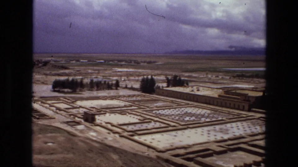 1972 Iran 8mm frame showing an elevated view across rectangular garden beds, low stone paths, scattered trees, and distant flat fields.