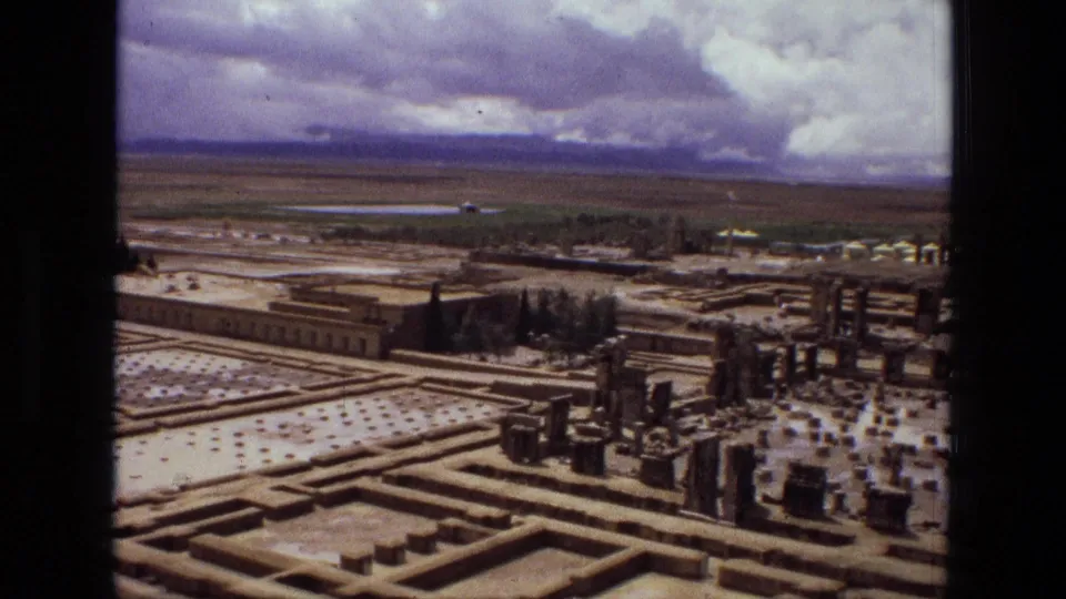 1972 Iran 8mm frame with geometric beds in the foreground, upright ruin-like stone features to the right, trees, and broad fields beyond.