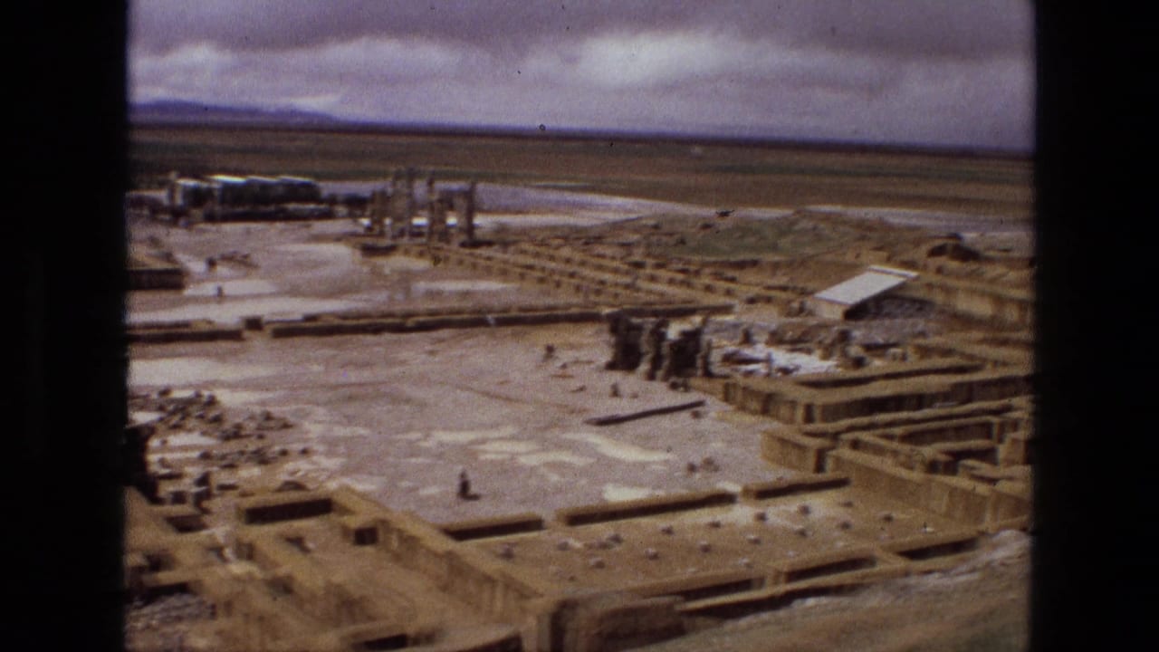 1972 Iran home-movie frame of an aerial pass over a ruin complex, with rectangular foundations, columns, and distant flats visible.