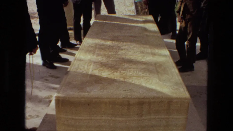 Iran 1972 funeral frame showing mourners in dark suits gathered around a carved stone tomb with inscriptions.