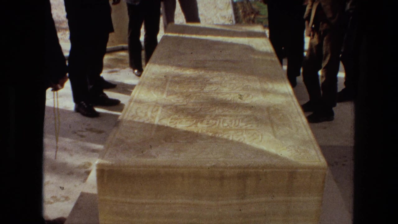 1972 Iran 8mm frame showing a carved stone sarcophagus with inscriptions, viewed close up among standing mourners in dark clothing.