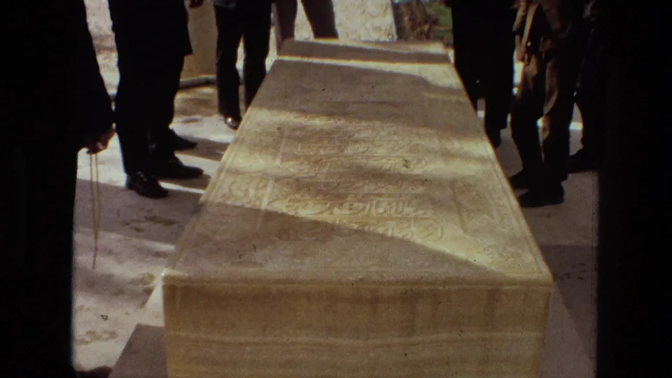 1972 Iran 8mm frame showing a carved stone sarcophagus with inscriptions, viewed close up among standing mourners in dark clothing.