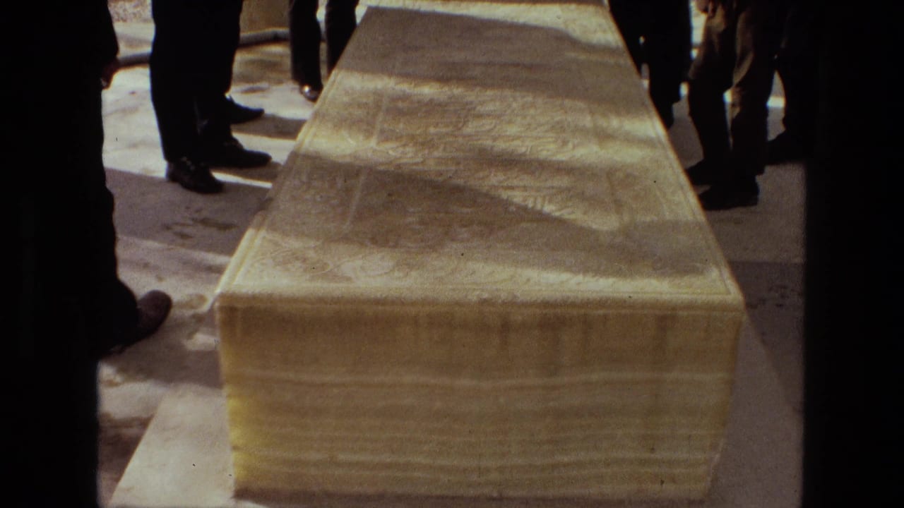 Close 1972 Iran home-movie view of a large stone sarcophagus with people gathered around its sides in a funeral or memorial setting.