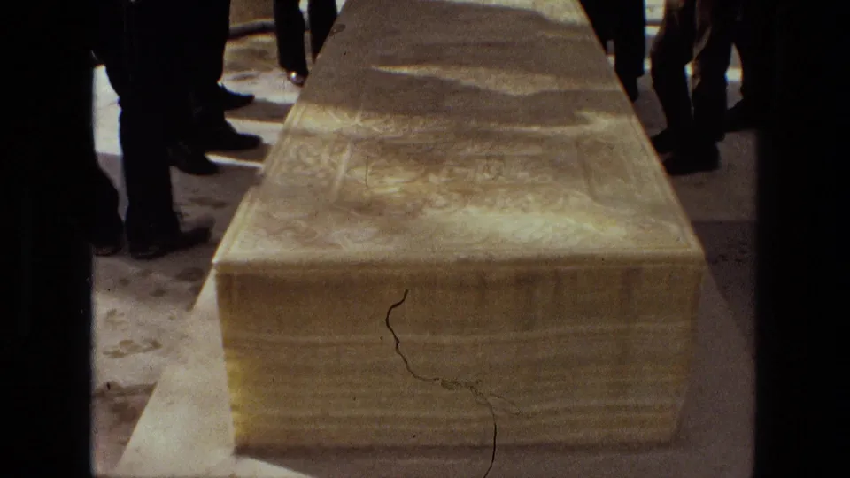 Handheld 8mm frame from 1972 Iran showing the top and end of an inscribed stone sarcophagus, with mourners partly visible around it.