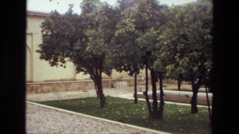 1972 Iran home-movie frame showing a calm courtyard with trees, grass, paving, and a traditional light-colored building in the background.