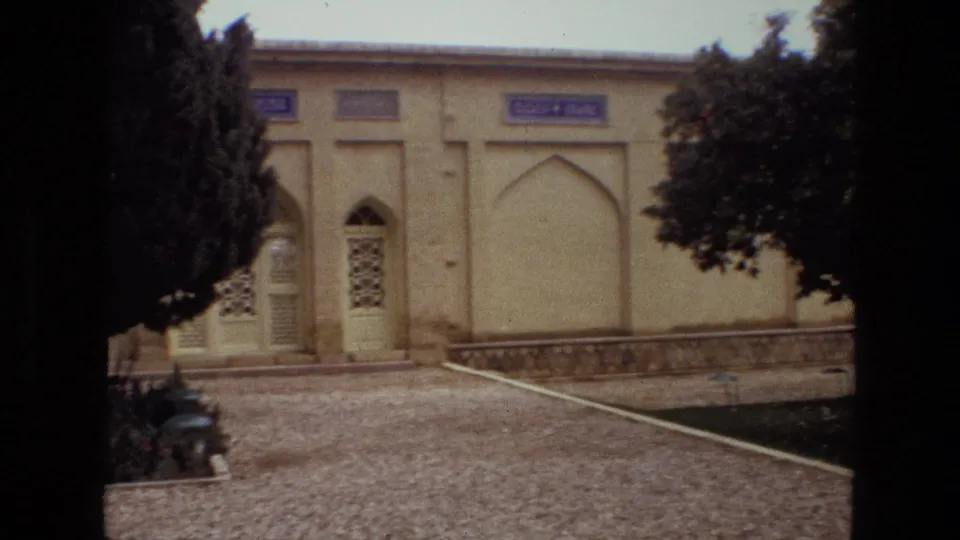 1972 Iran 8mm frame with arched doorways, decorative panels, gravel paths and trees in a landscaped courtyard.